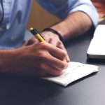Une main écrit sur du papier avec un stylo, un ordinateur portable à côté