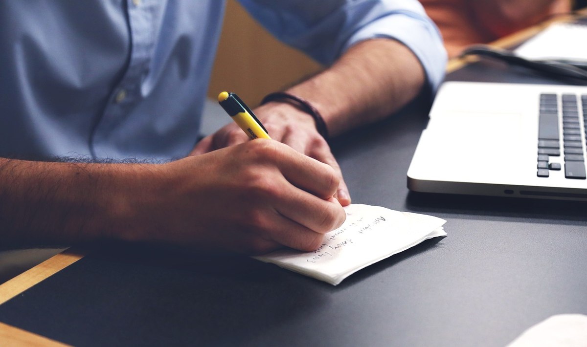 Une main écrit avec un stylo sur un papier, à côté d'un ordinateur